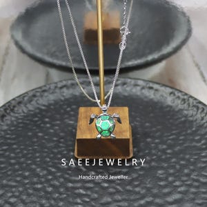 May include: A silver necklace with a turtle pendant. The pendant features a green, faceted center and silver detailing. The necklace is displayed on a wooden stand against a dark background. The text "SAEEJEWELRY" and "Handcrafted Jeweller" are visible.