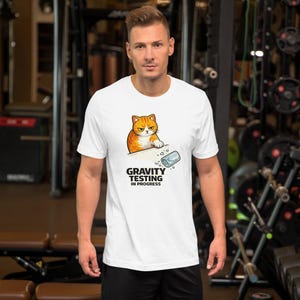 Può includere: T-shirt bianca con un gatto tabby arancione dei cartoni animati con un'espressione accigliata. Il gatto è appoggiato su una superficie con un bicchiere rovesciato e la scritta "GRAVITY TESTING IN PROGRESS".