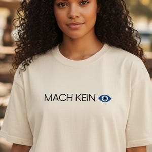 以下が含まれることがあります： 「MACH KEIN」の文字とネイビーブルーの目のグラフィックが入ったクリーム色のTシャツ。半袖でゆったりとしたフィット感。モデルは屋外でTシャツを着ています。