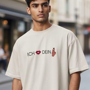 Könnte beinhalten: Beiges T-Shirt mit dem deutschen Text "ICH 💋 DEIN" und einer herzförmigen Handgeste. Das Shirt hat kurze Ärmel und eine lockere Passform. Das Model steht draußen auf einer Stadtstraße.