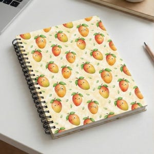 May include: Spiral-bound notebook featuring a repeating pattern of cartoon mangoes with faces and green leaves on a cream background. The notebook has a black spiral binding and is open on a white surface.