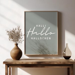 Puede incluir: Un cuadro con la palabra "Hallo" en escritura blanca, junto con "Halli" y "Hallöchen" en un diseño minimalista. La impresión se muestra sobre una mesa de madera, acompañada de un jarrón con flores secas y una botella de vidrio decorativa.