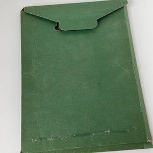 May include: A green paper envelope with a flap closure. The envelope is rectangular and has a textured surface. The flap has a curved design with a small cutout. The envelope is slightly worn.