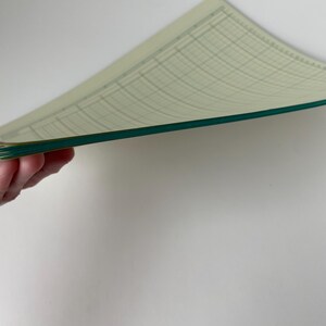 May include: A stack of graph paper sheets with a light green grid pattern and a dark green border. The paper is slightly curved, revealing the edges and the grid lines. The background is a plain white surface.