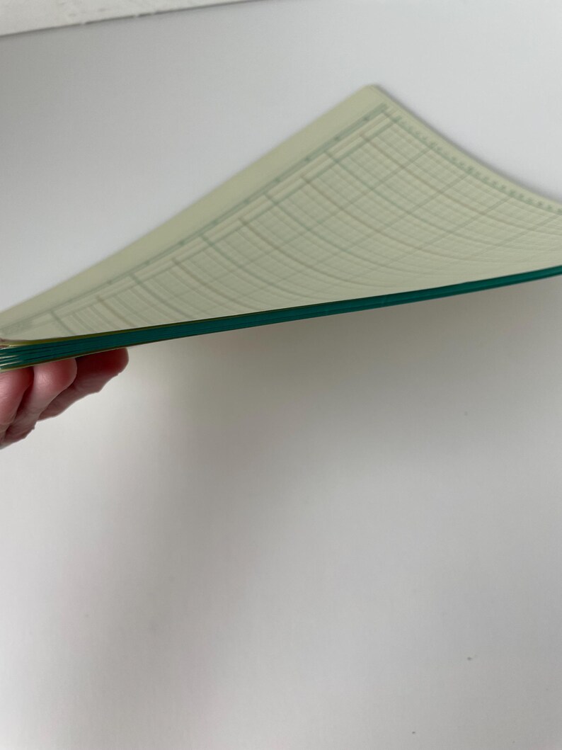 May include: A stack of graph paper sheets with a light green grid pattern and a dark green border. The paper is slightly curved, revealing the edges and the grid lines. The background is a plain white surface.