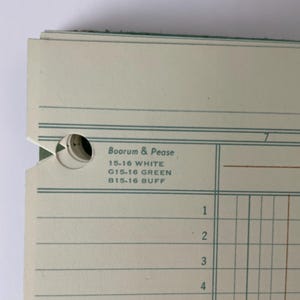 May include: Close-up of a stack of ledger paper with a green border. The paper has horizontal and vertical lines, creating a grid. Text includes "Boorum & Pease" and color codes: "15-16 WHITE", "G15-16 GREEN", and "B15-16 BUFF".