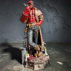 Può includere: Una statuetta umanoide dalla pelle rossa con corna, pizzetto e una grande mano destra, che indossa un trench marrone, pantaloni neri e stivali marroni. La figura poggia su una base a tema cimiteriale con un teschio e delle candele.