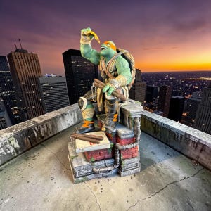 Puede incluir: Estatua de Miguel Ángel de las Tortugas Ninja, sosteniendo una porción de pizza, en una azotea con un fondo de paisaje urbano. La estatua es verde, naranja y marrón, con un caparazón y armas detalladas. También se ve una caja de pizza y un paisaje urbano.