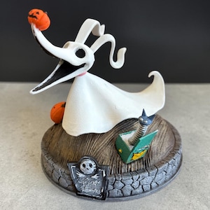 Może przedstawiać: Figurka Zero, psa ducha z Miasteczka Halloween, trzymającego pomarańczową dynię. Biała postać ma długi, zakrzywiony nos i mały różowy kołnierz. Stoi na drewnianej podstawie z nagrobkiem i książką.