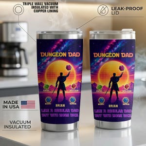 May include: Two stainless steel vacuum-insulated tumblers with a colorful design. The design features the text "DUNGEON DAD" above a silhouette of a figure holding a die, with names below. The tumblers have a leak-proof lid.