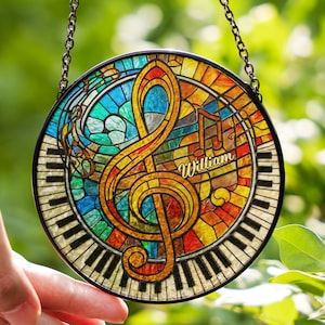 May include: A round, stained-glass suncatcher featuring a musical theme. The design includes a large treble clef, musical notes, and piano keys. The name "William" is written in cursive. The suncatcher is framed with a black border and hangs from a chain.