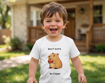May include: A white short-sleeved t-shirt featuring a cartoon illustration of a brown capybara holding a red apple. The text "Don't worry" is printed above the capybara, and the text "Be Cappy" is printed below the illustration in black.