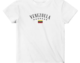 May include: White t-shirt featuring the word "VENEZUELA" in black arched text above a small Venezuelan flag graphic. The design includes six small black stars. A simple, patriotic design.