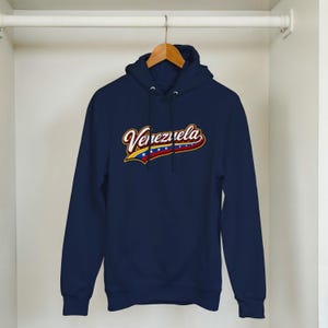 May include: A navy blue hooded sweatshirt featuring the word Venezuela in a stylized white and red script font. Below the text, a graphic element incorporates the yellow, blue, and red colors of the Venezuelan flag with a row of white stars.