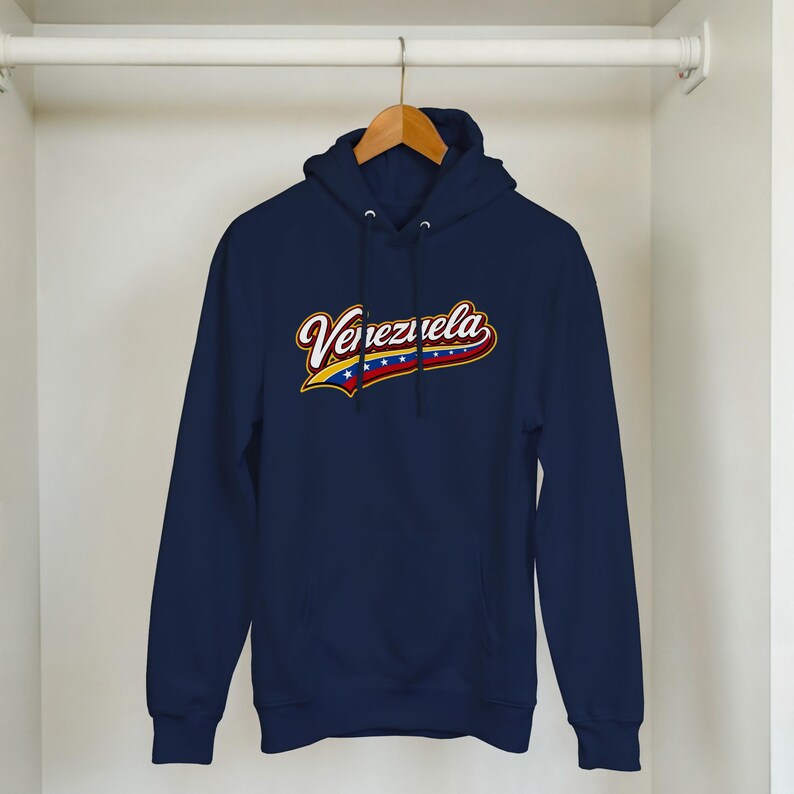 May include: A navy blue hooded sweatshirt featuring the word Venezuela in a stylized white and red script font. Below the text, a graphic element incorporates the yellow, blue, and red colors of the Venezuelan flag with a row of white stars.