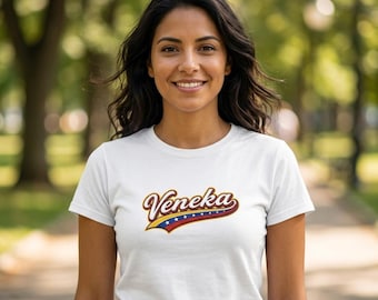 May include: A white short-sleeved t-shirt featuring the word Veneka in a stylized script font. The text is outlined in dark brown and sits above a curved graphic element colored in yellow, blue, and red with a row of small white stars.