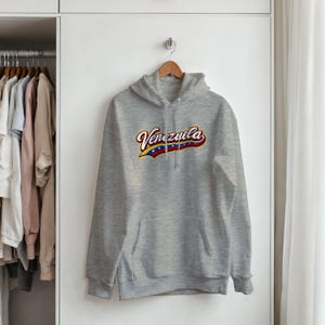May include: A light gray hooded sweatshirt featuring the word Venezuela printed across the chest in a stylized font. The text incorporates the yellow, blue, and red colors of the Venezuelan flag with white stars along the bottom edge.