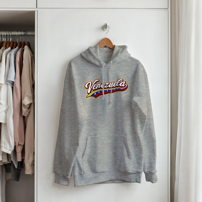 May include: A light gray hooded sweatshirt featuring the word Venezuela printed across the chest in a stylized font. The text incorporates the yellow, blue, and red colors of the Venezuelan flag with white stars along the bottom edge.