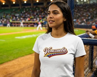May include: A white short-sleeved t-shirt featuring the word Venezuela in a stylized script font. The text is outlined in dark blue and gold, with a graphic element below the lettering that displays the yellow, blue, and red colors of the flag.