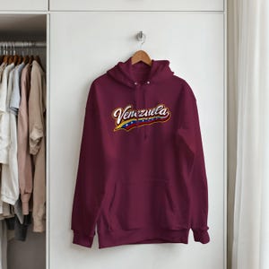 May include: A maroon long-sleeve hooded sweatshirt featuring a front kangaroo pocket and a graphic across the chest that reads Venezuela in white script with a yellow, blue, and red flag-inspired underline containing small white stars.