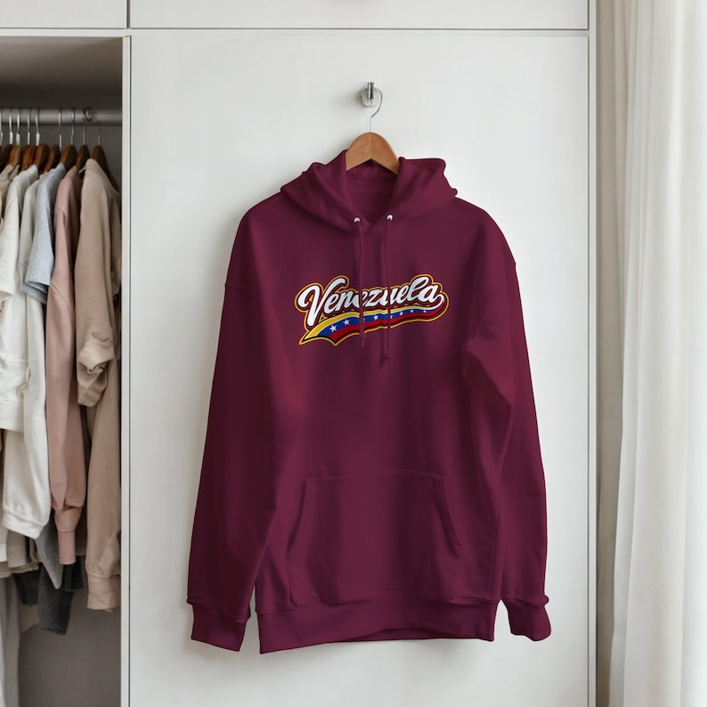 May include: A maroon long-sleeve hooded sweatshirt featuring a front kangaroo pocket and a graphic across the chest that reads Venezuela in white script with a yellow, blue, and red flag-inspired underline containing small white stars.