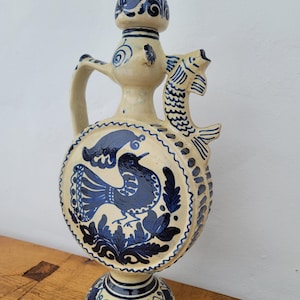 May include: A ceramic pitcher with a cream-colored base and intricate blue designs. The pitcher features a bird and floral motif, a fish-shaped handle, and a decorative top. The base is round and supported by a pedestal. The pitcher is approximately 12 inches tall.