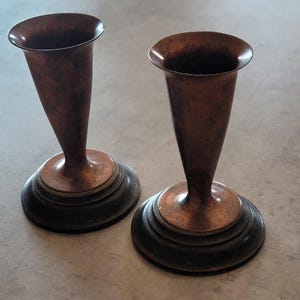 Könnte beinhalten: Ein Paar passende Kerzenhalter mit einem ausgestellten, trompetenförmigen Oberteil aus Kupfer mit einer verwitterten Patina. Jeder Halter wird von einem breiten, kreisförmigen, dunkel getönten Sockel getragen, der dem Metallschaft Halt bietet.
