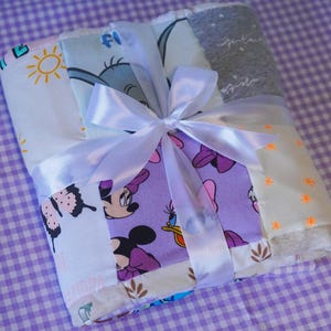 May include: A folded baby blanket tied with a white satin ribbon. The blanket features various colorful patterned fabrics, including Minnie Mouse, butterflies, and suns. The blanket is on a purple and white checkered surface.