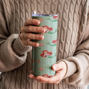May include: A green tumbler featuring a repeating pattern of a mermaid with red hair and a green tail, seashells, and seaweed. The tumbler has a silver lid and a white straw. The background is a light green color.