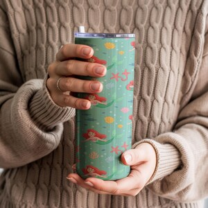 May include: A teal tumbler with a mermaid-themed design. The tumbler features illustrations of mermaids, starfish, seashells, and bubbles. The tumbler has a silver lid and a white straw.