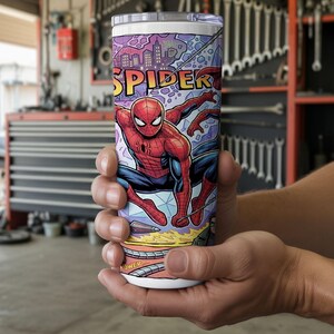 May include: A cylindrical stainless steel tumbler featuring a colorful comic book graphic of a red and blue costumed superhero in a dynamic pose. The design includes the word SPIDER in bold yellow letters against a purple city skyline background.