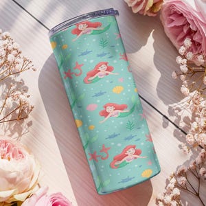 May include: A teal tumbler featuring a mermaid pattern with red-haired mermaids, starfish, seashells, and seaweed. The tumbler is cylindrical and has a clear lid. The design is set against a light teal background. The tumbler is surrounded by pink flowers and white flowers.