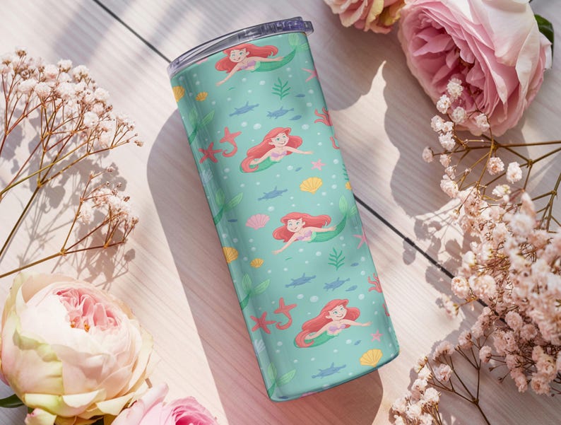 May include: A teal tumbler featuring a mermaid pattern with red-haired mermaids, starfish, seashells, and seaweed. The tumbler is cylindrical and has a clear lid. The design is set against a light teal background. The tumbler is surrounded by pink flowers and white flowers.