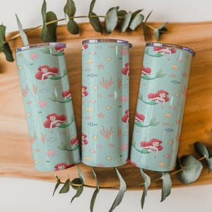 May include: Three teal insulated tumblers with a mermaid pattern. Each tumbler features a repeating design of a red-haired mermaid, seashells, fish, and coral. The tumblers are cylindrical and have a silver lid. The background is a wooden surface with green leaves.