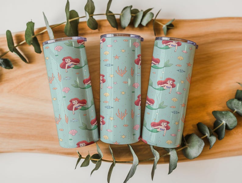 May include: Three teal insulated tumblers with a mermaid pattern. Each tumbler features a repeating design of a red-haired mermaid, seashells, fish, and coral. The tumblers are cylindrical and have a silver lid. The background is a wooden surface with green leaves.