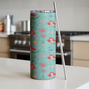 May include: A teal tumbler with a mermaid pattern, featuring red-haired mermaids, coral, starfish, and fish. Includes a silver straw. The tumbler is cylindrical and stands on a white surface.