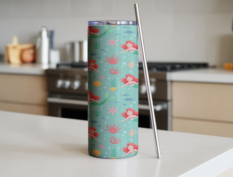 May include: A teal tumbler with a mermaid pattern, featuring red-haired mermaids, coral, starfish, and fish. Includes a silver straw. The tumbler is cylindrical and stands on a white surface.