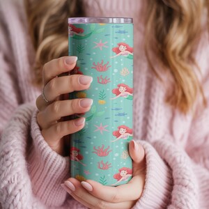May include: A teal tumbler featuring a repeating pattern of a mermaid with red hair, swimming among coral, starfish, and fish. The tumbler has a silver rim and a clear lid. The design is whimsical and colorful.