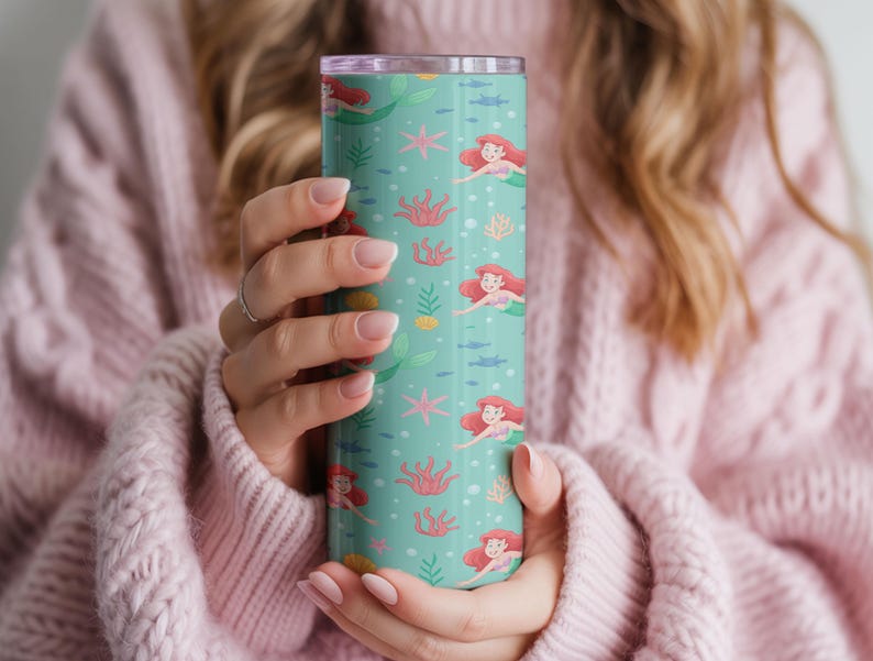 May include: A teal tumbler featuring a repeating pattern of a mermaid with red hair, swimming among coral, starfish, and fish. The tumbler has a silver rim and a clear lid. The design is whimsical and colorful.