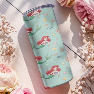 May include: A teal tumbler featuring a repeating pattern of a mermaid with red hair and a green tail, along with seashells, fish, and starfish. The tumbler has a clear lid and is surrounded by pink flowers and white flowers.