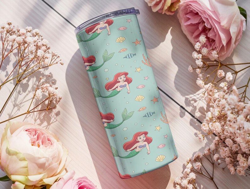 May include: A teal tumbler featuring a repeating pattern of a mermaid with red hair and a green tail, along with seashells, fish, and starfish. The tumbler has a clear lid and is surrounded by pink flowers and white flowers.
