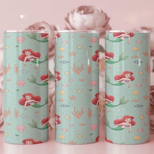 May include: Three teal-colored tumblers with a repeating pattern of a mermaid, seashells, fish, and seaweed. The mermaid has red hair and a green tail. The tumblers are cylindrical and have a silver lid.