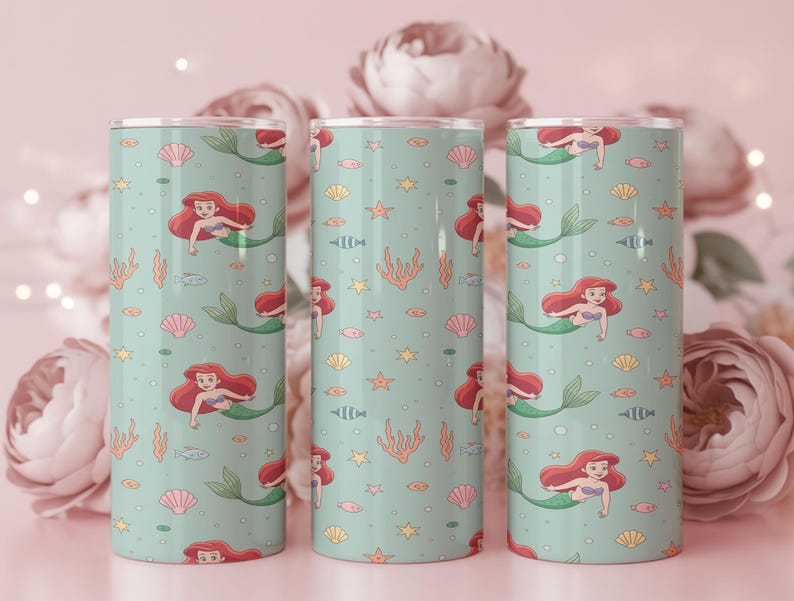 May include: Three teal-colored tumblers with a repeating pattern of a mermaid, seashells, fish, and seaweed. The mermaid has red hair and a green tail. The tumblers are cylindrical and have a silver lid.