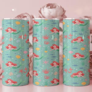 May include: Three cylindrical tumblers with a teal background featuring a pattern of mermaids, seashells, starfish, and seaweed. The mermaids have red hair and green tails. The tumblers are stainless steel.