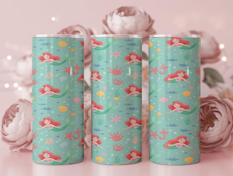 May include: Three cylindrical tumblers with a teal background featuring a pattern of mermaids, seashells, starfish, and seaweed. The mermaids have red hair and green tails. The tumblers are stainless steel.