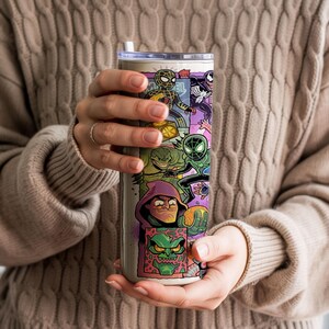 May include: A stainless steel tumbler with a vibrant comic book character design. The artwork includes Spider-Man, Venom, and other heroes and villains. The tumbler has a white straw. The person holding the tumbler is wearing a beige jumper.