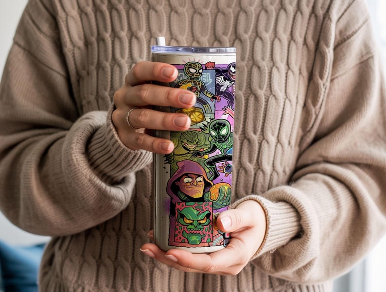 May include: A stainless steel tumbler with a vibrant comic book character design. The artwork includes Spider-Man, Venom, and other heroes and villains. The tumbler has a white straw. The person holding the tumbler is wearing a beige jumper.