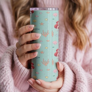 May include: A teal tumbler featuring a repeating pattern of red-haired mermaids, seashells, starfish, and coral. The tumbler is cylindrical and has a silver lid. The design is whimsical and evokes a nautical theme.