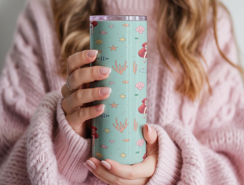 May include: A teal tumbler featuring a repeating pattern of red-haired mermaids, seashells, starfish, and coral. The tumbler is cylindrical and has a silver lid. The design is whimsical and evokes a nautical theme.