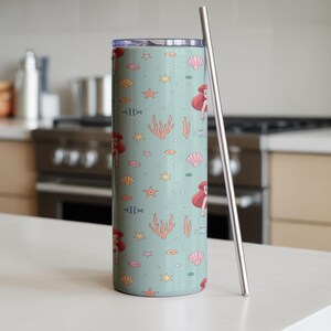 May include: A tall, cylindrical tumbler with a light teal background featuring a repeating pattern of a mermaid, seashells, starfish, coral, and fish. A silver-colored straw rests against the side of the tumbler. The tumbler is designed for cold beverages.